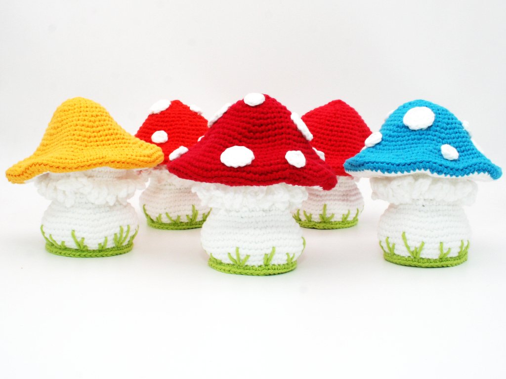 Five crocheted mushroom-shaped egg cosies with red, blue and yellow caps, white ruffled stems and green grass-edged bases
