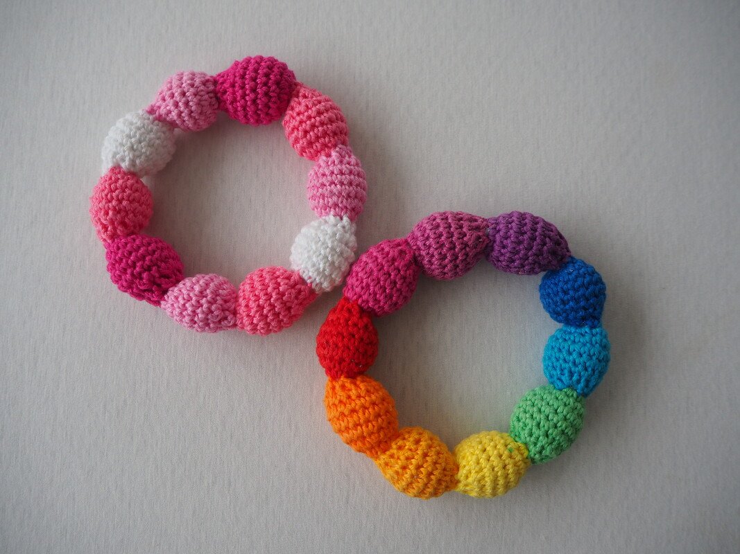 Zwei gehäkelte Baby-Greifringe auf hellem Untergrund, einer rosa-weiß, der andere regenbogenfarben