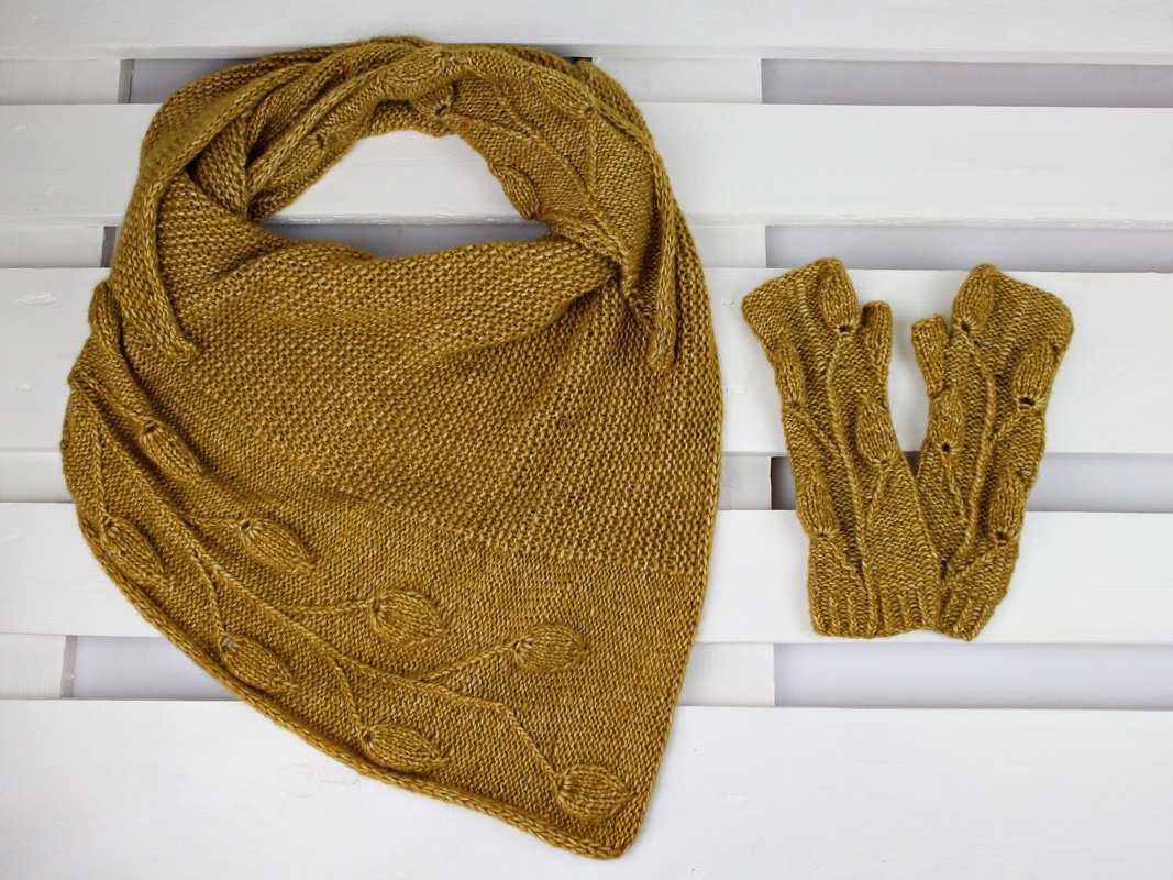 Gestricktes senfgelbes Dreieckstuch und fingerlose Handschuhe mit Blättermuster auf weißem Holzuntergrund.