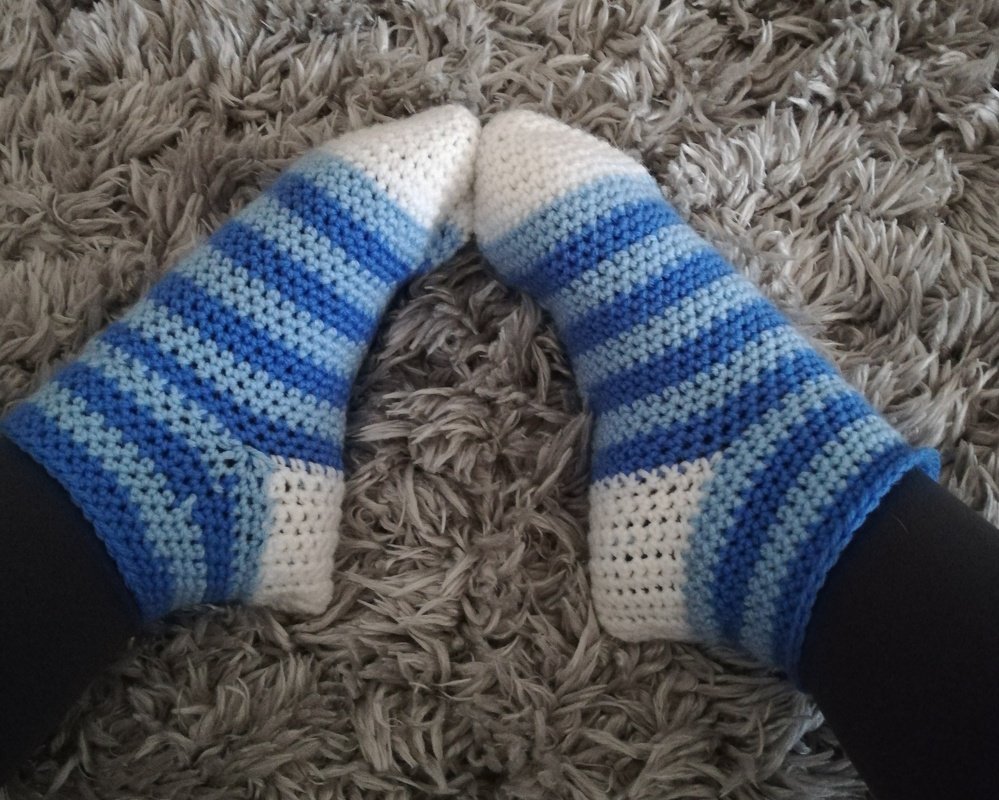 Paar Füße in blau-weiß gestreiften, gehäkelten Socken auf einem grauen, flauschigen Teppich.