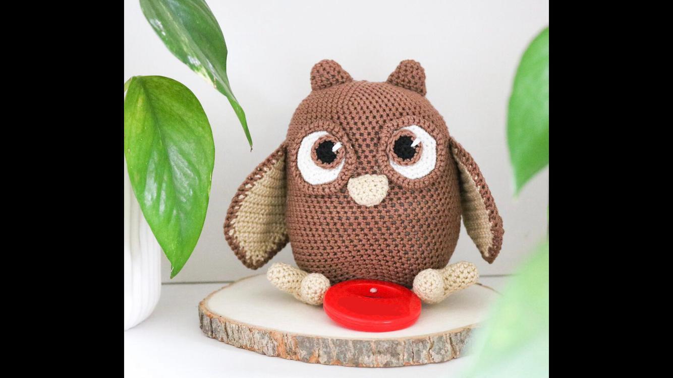 Gehäkelte Amigurumi-Eule (braun/beige) sitzend auf Holzscheibe, kleines rotes Objekt vorne, grüne Blätter im Hintergrund