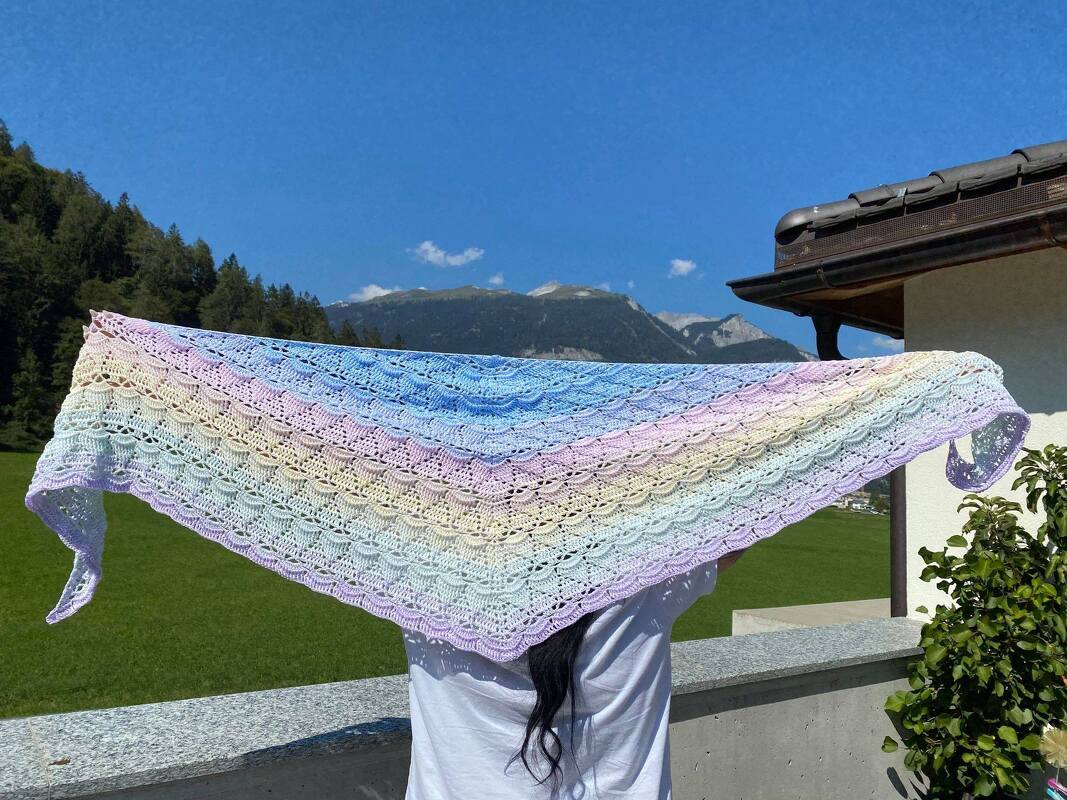Person hält großes gehäkeltes Dreieckstuch in Pastellfarben mit Lochmuster vor Bergkulisse
