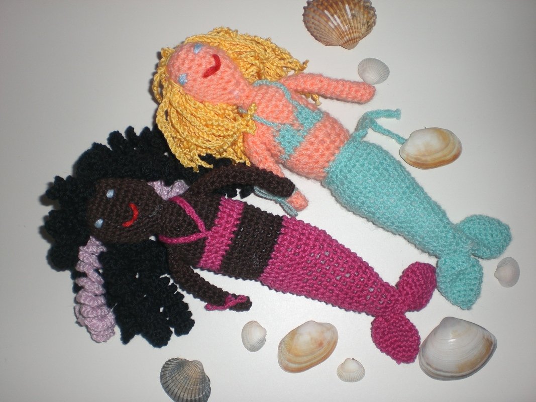 Zwei gehäkelte Amigurumi-Meerjungfrauenpuppen mit unterschiedlichen Haar- und Schwanzfarben zwischen Muscheln.