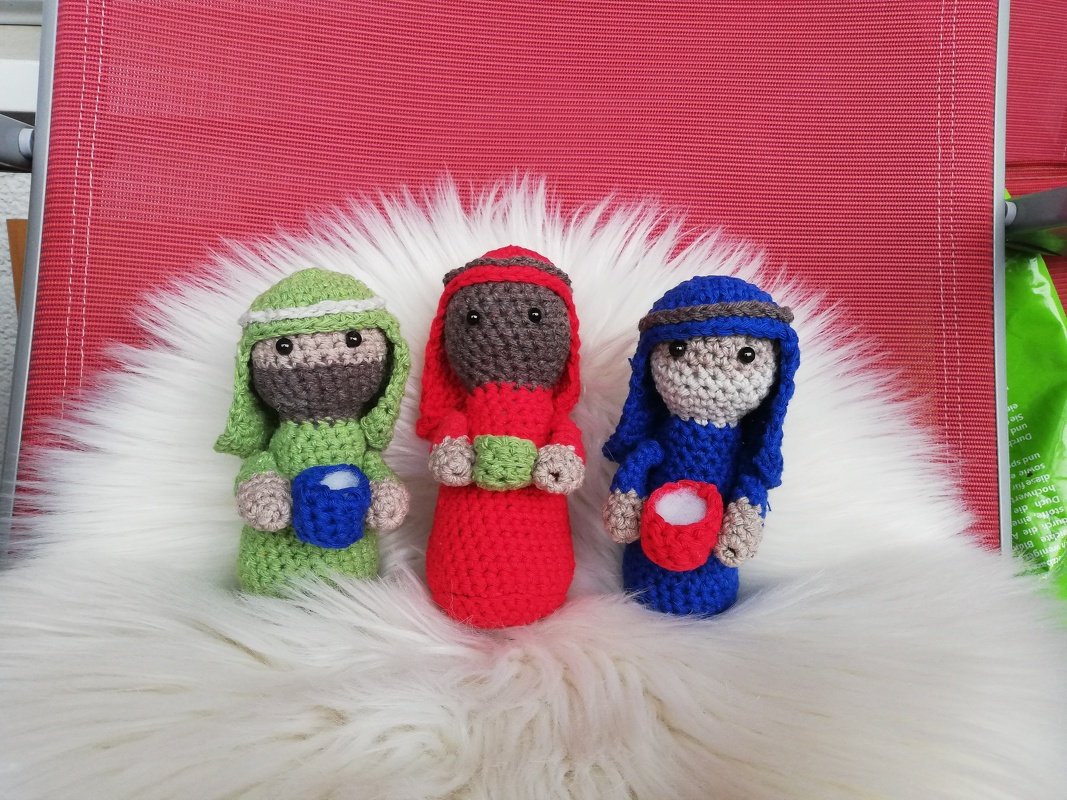 Drei gehäkelte Amigurumi-Könige in Grün, Rot und Blau auf weißem Kunstfell vor rotem Hintergrund.