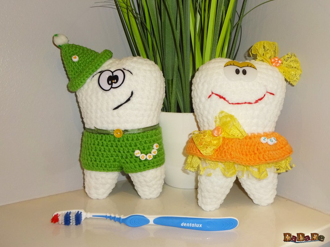 Zahn-Amigurumi-Duo mit Häkelmütze, Hose, Rock und Schleifen neben einer blauen Zahnbürste