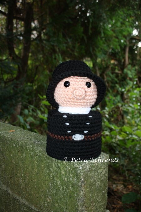 Gehäkelte Schornsteinfeger-Figur in schwarzer Kleidung auf moosbewachsener Steinmauer vor grünem Hintergrund