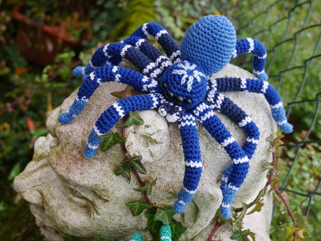 Blau-weiß gestreifte Amigurumi-Häkelspinne auf verwittertem Steinkopf im Garten.