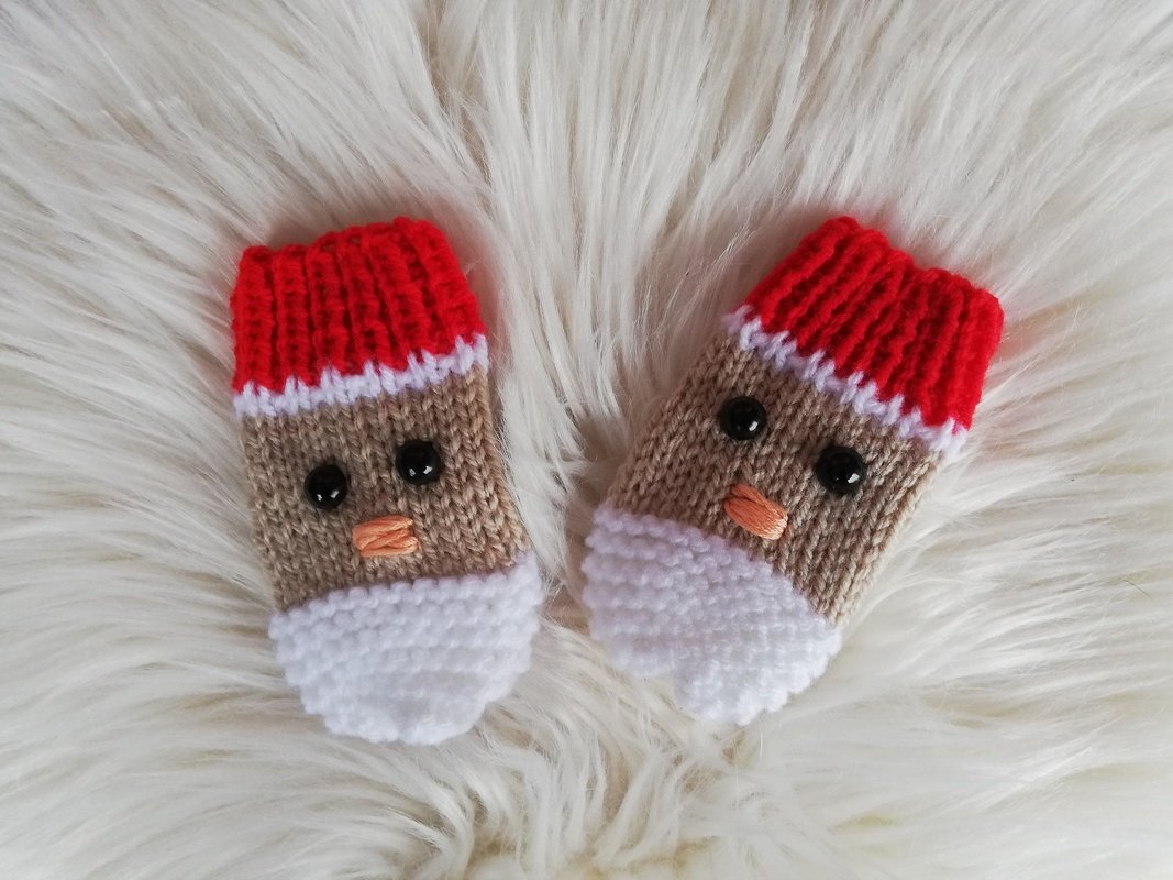 Zwei handgestrickte Baby‑Fäustlinge in Rot, Beige und Weiß mit schwarzen Knopfaugen und gestickter oranger Nase auf flauschigem, hellem Hintergrund