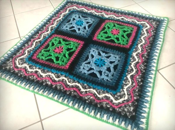 Bunt gehäkelte quadratische Decke mit vier Ornamentquadraten auf hellem Fliesenboden.