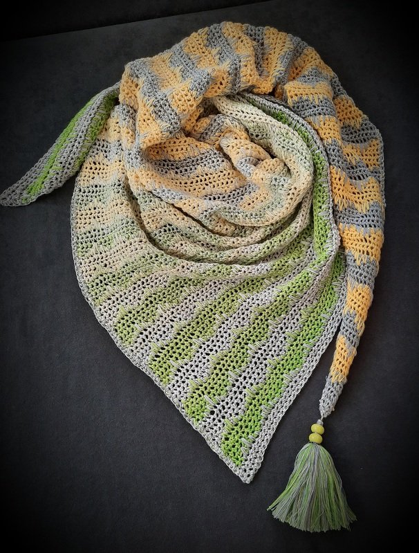 Gehäkeltes Dreieckstuch in Gelb, Grau und Hellgrün auf dunklem Hintergrund. Das Tuch zeigt Streifen- und Lochmuster sowie eine Quaste mit Perlen an einer Spitze.