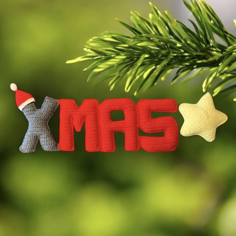 Gehäkelte Buchstaben 'XMAS' in Rot und Grau mit kleinem gelben Stern vor grünem Hintergrund