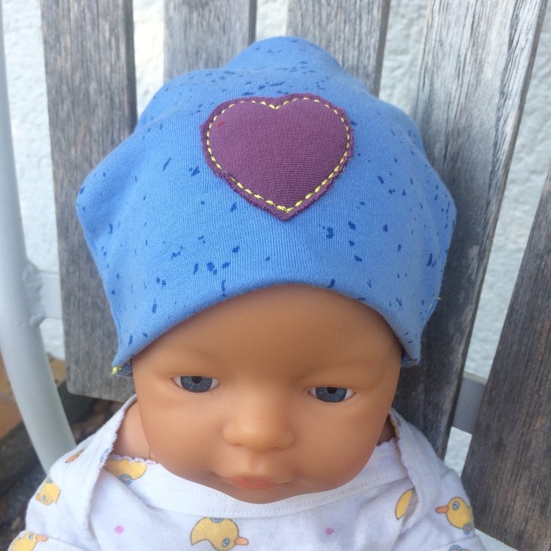 Babypuppe mit blauem Mützchen und lila Herz-Applikation mit gelber Ziernaht