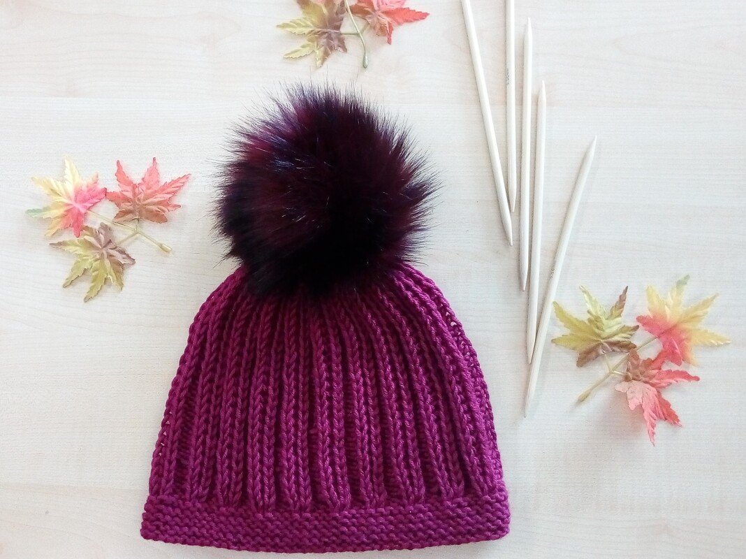 Lila Rippenstrickmütze mit dunklem Bommel auf hellem Holzuntergrund, daneben Stricknadeln und Herbstblätter.