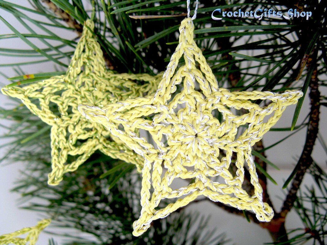 Two pale yellow crocheted star ornaments with silver metallic thread hanging on pine needles.