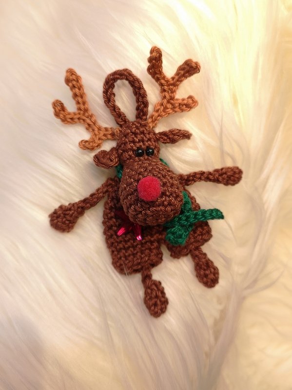 Gehäkeltes braunes Rentier mit roter Nase, grünem Schal und Aufhängeschlaufe auf hellem Fellhintergrund