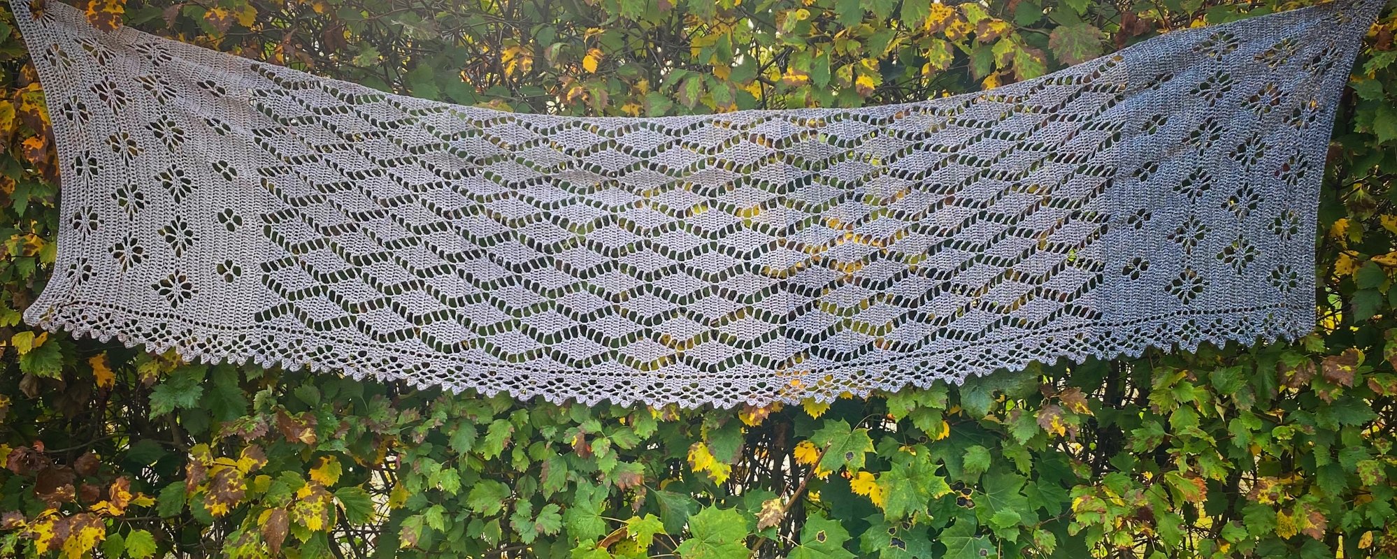 Light gray rectangular crochet shawl with diamond and floral lace motifs hanging against green and yellow leaves.