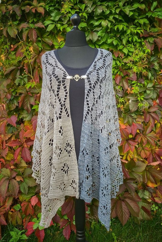 Two-tone grey to blue-grey crochet rectangle shawl with openwork lace and floral motifs, fastened with an ornamental metal clasp on a black mannequin.