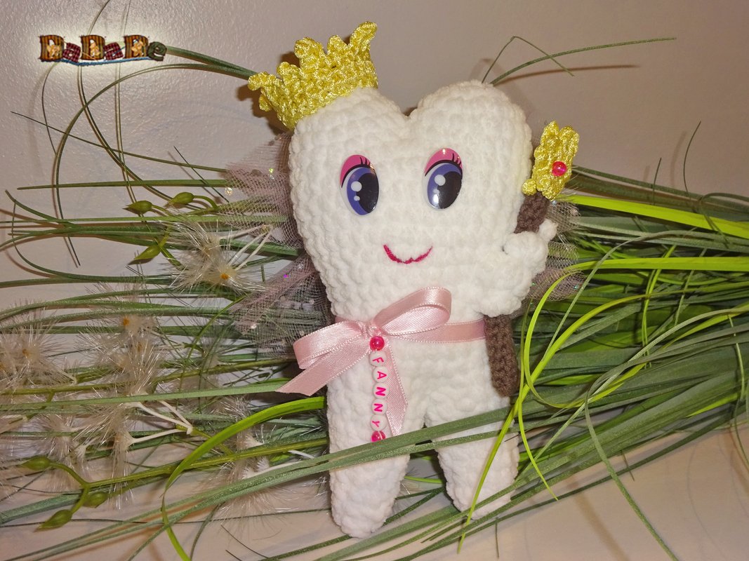 Gehäkelte weiße Zahnfee mit goldener Krone, rosa Schleife, Zauberstab und Flügeln vor dekorativen Gräsern