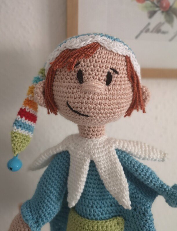 Gehäkelte Amigurumi-Puppe mit orangebraunem Haar, blauem Oberteil und gestreifter Zipfelmütze mit blauer Perle