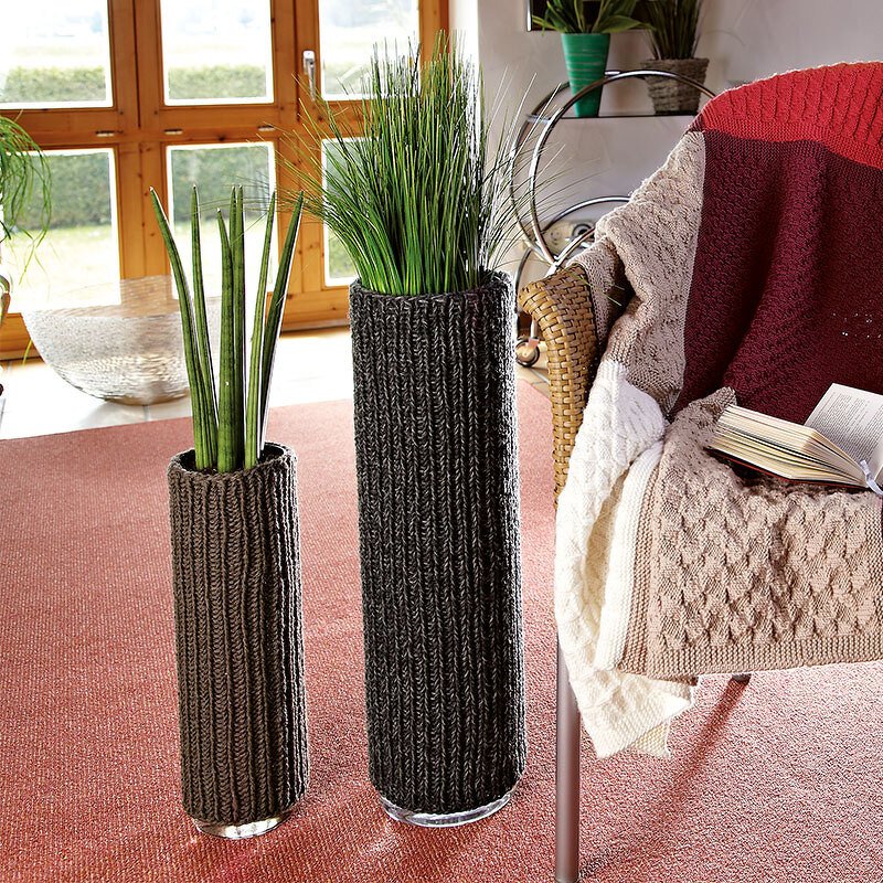Zwei hohe Glasvasen mit dunkelgrauen gestrickten Überzügen auf rotem Teppich neben einem Sessel mit Strickdecke