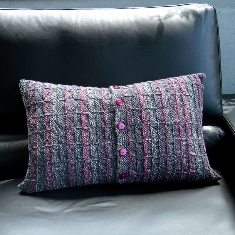 Gestricktes rechteckiges Kissen grau-rosa mit drei pinken Knöpfen auf schwarzer Couch