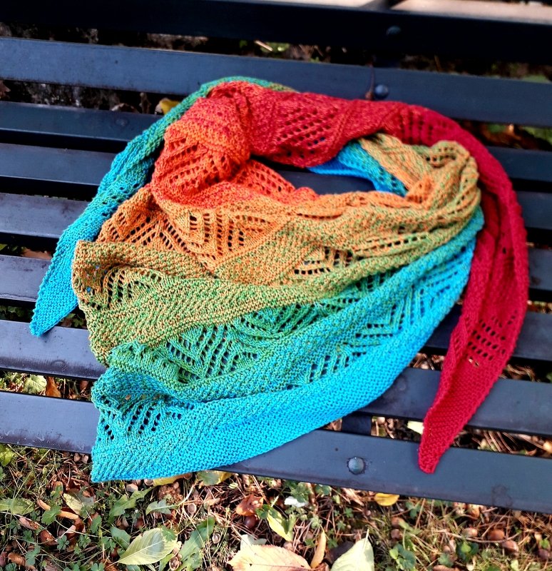 Gestricktes, buntes Dreieckstuch mit Ajourmuster und sanftem Farbverlauf, auf einer Parkbank drapiert
