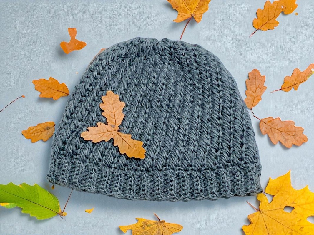 Gehäkelte graue Beanie auf hellem Hintergrund, umgeben von herbstlichen Blättern