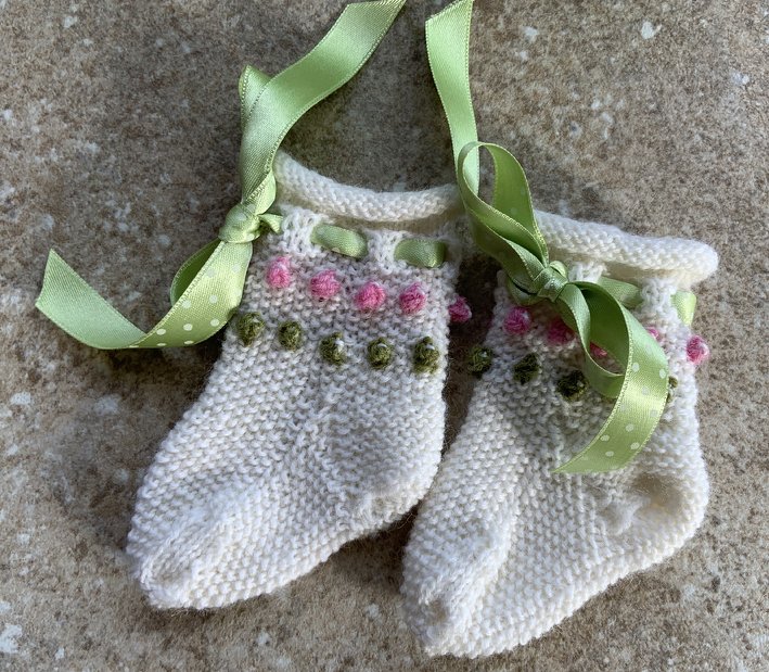 Zwei cremefarbene, gestrickte Babysocken mit rosa und olivgrünen Noppen und grünen Satinbändern, eines gepunktet.