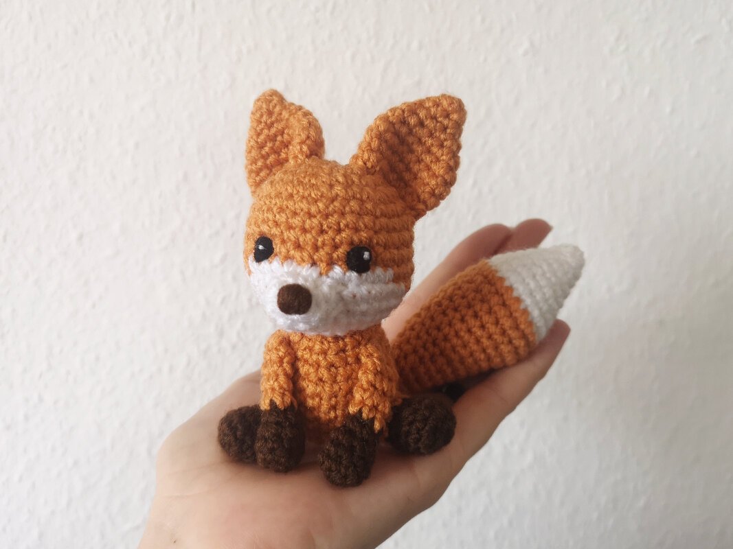 Hand holding a small orange and white crocheted fox amigurumi with brown feet and a tapered tail