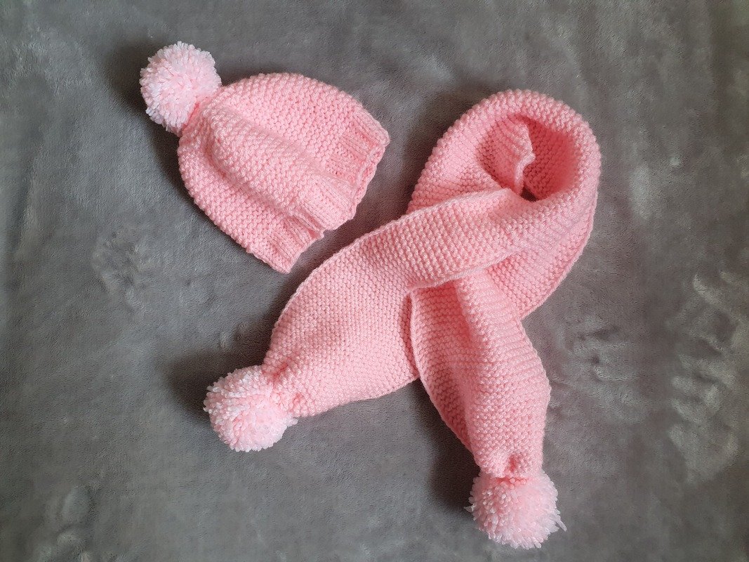 Rosa gestrickte Babymütze mit Bommel und passender Schal mit Bommeln auf grauem Hintergrund