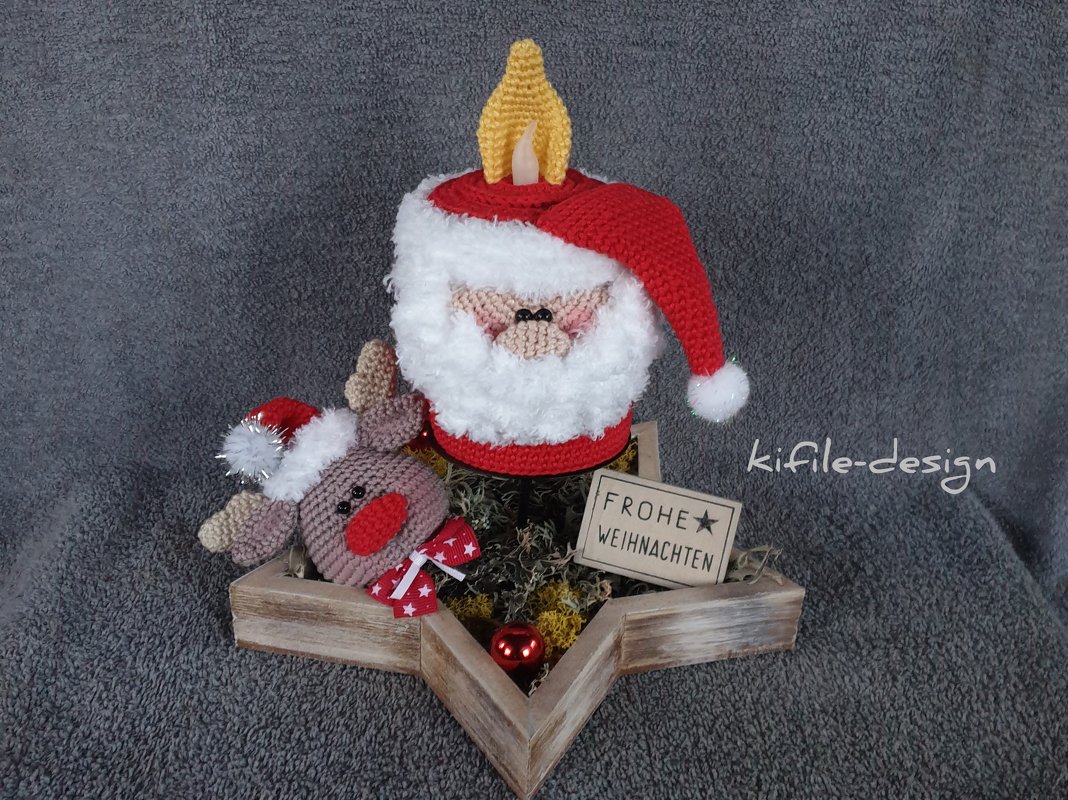 Gehäkelte Weihnachtsmann-Kerzenhülle mit gehäkelter Flamme und ein gehäkeltes Rentier mit roter Schleife in einer sternförmigen Holzschale mit Moos und Schild ‚Frohe Weihnachten‘.