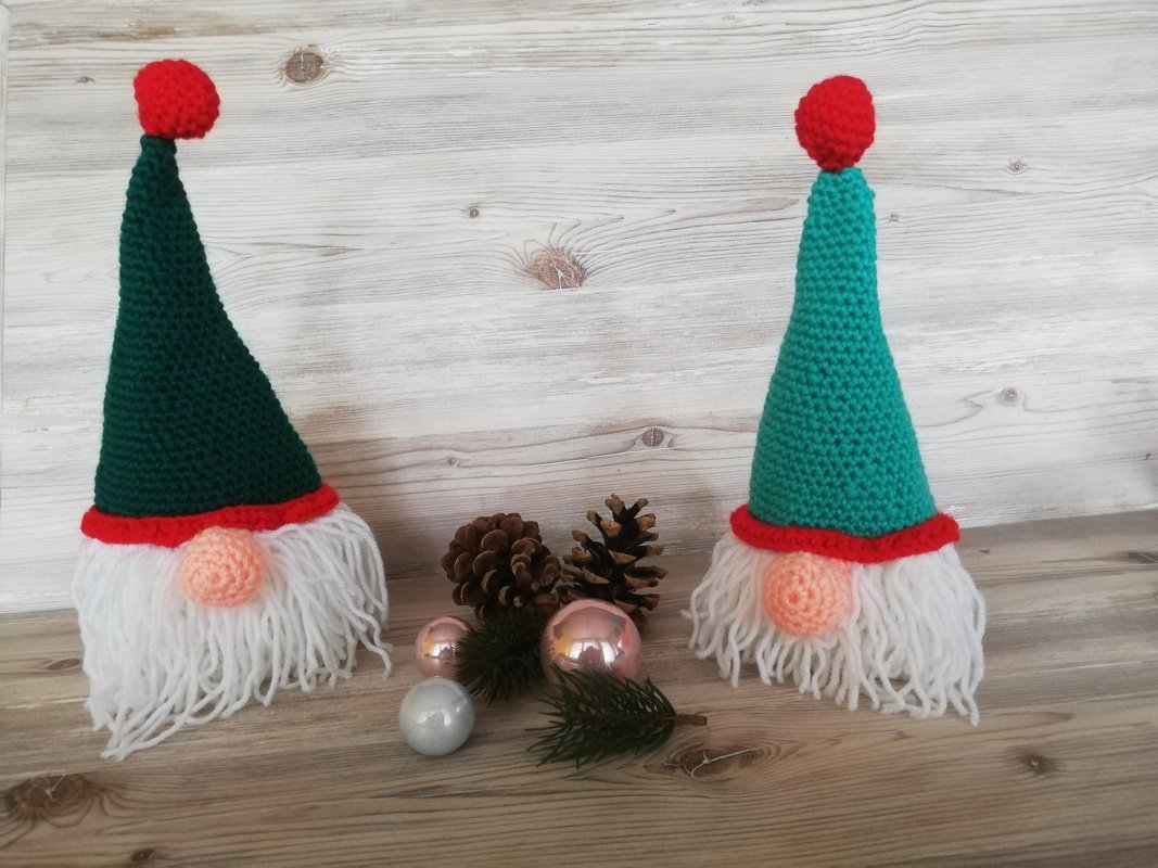 Zwei gehäkelte Wichtel mit weißen Bärten, grüner und türkiser Mütze und roten Bommeln, umgeben von Tannenzapfen und Weihnachtskugeln.