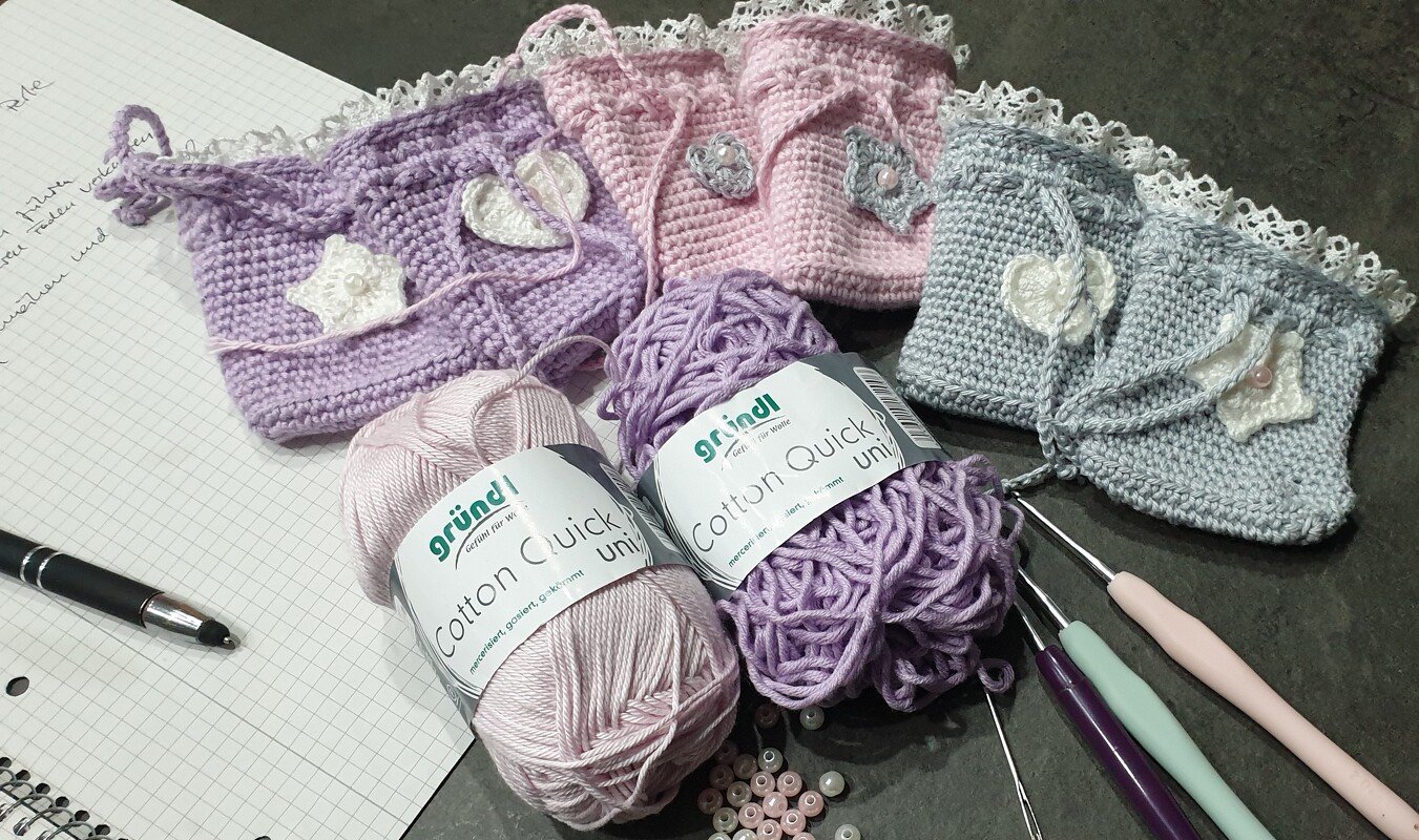 Drei gehäkelte Pastellbeutel mit Garnknäueln, Häkelnadeln und Perlen auf einer Arbeitsfläche.