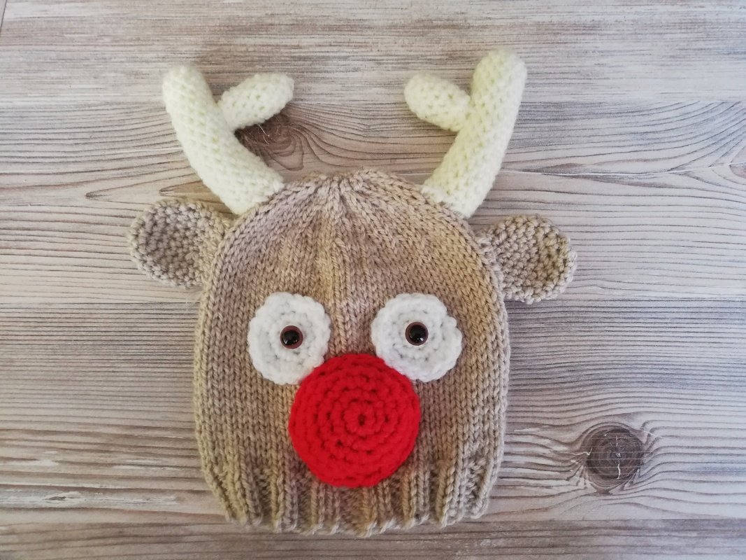 Beige-braune gestrickte Babymütze mit Rentier-Motiv, weißen Augen, kleinen Ohren, weißen Geweih-Applikationen und großer roter Nase auf Holzuntergrund.