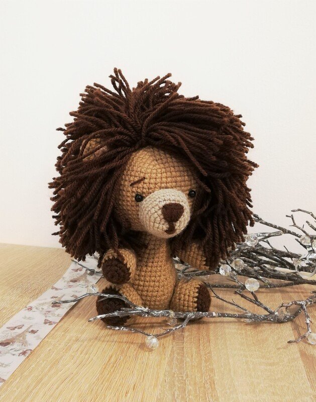 Gehäkelter Amigurumi-Löwe mit zotteliger brauner Mähne, sitzend auf einem Holztisch neben Dekozweigen.
