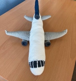 Gehäkeltes Amigurumi‑Flugzeug in Weiß und Grau auf Holztisch, Draufsicht