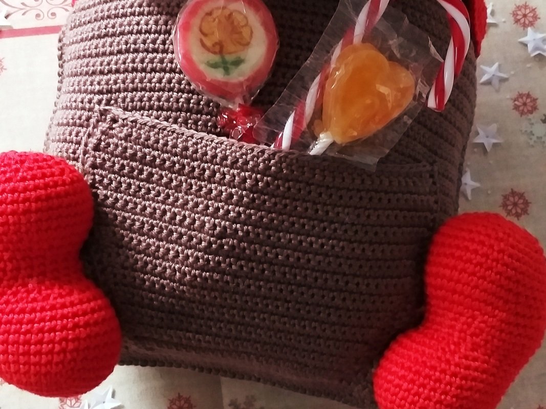 Brown crocheted character with red accents and a front pocket holding lollipops and a candy cane on a holiday-themed background.