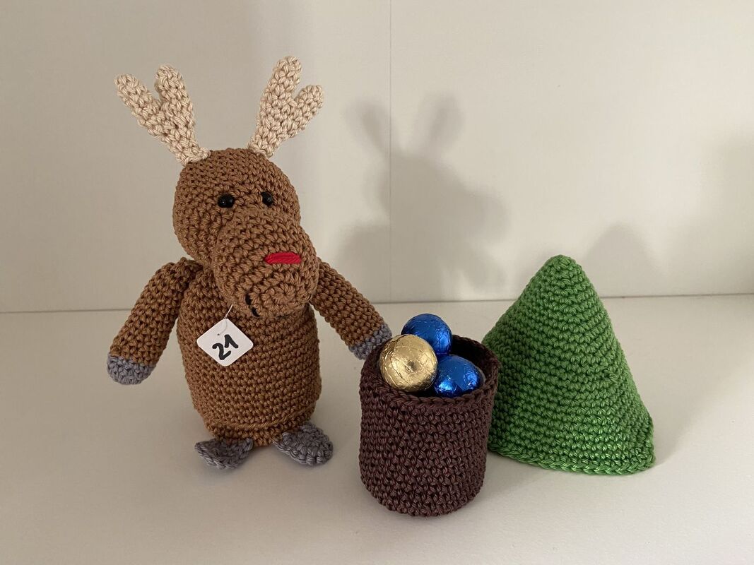 Gehäkeltes Rentier mit Schild „21“ neben Häkelkorb mit bunten Kugeln und grünem Häkelbaum.