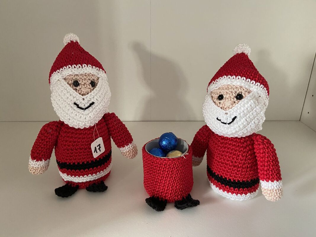 Gehäkelter Weihnachtsmann mit Zahlenanhänger 17 neben einem roten Häkelsäckchen mit Schokokugeln sowie einer zweiten Weihnachtsmann-Figur.