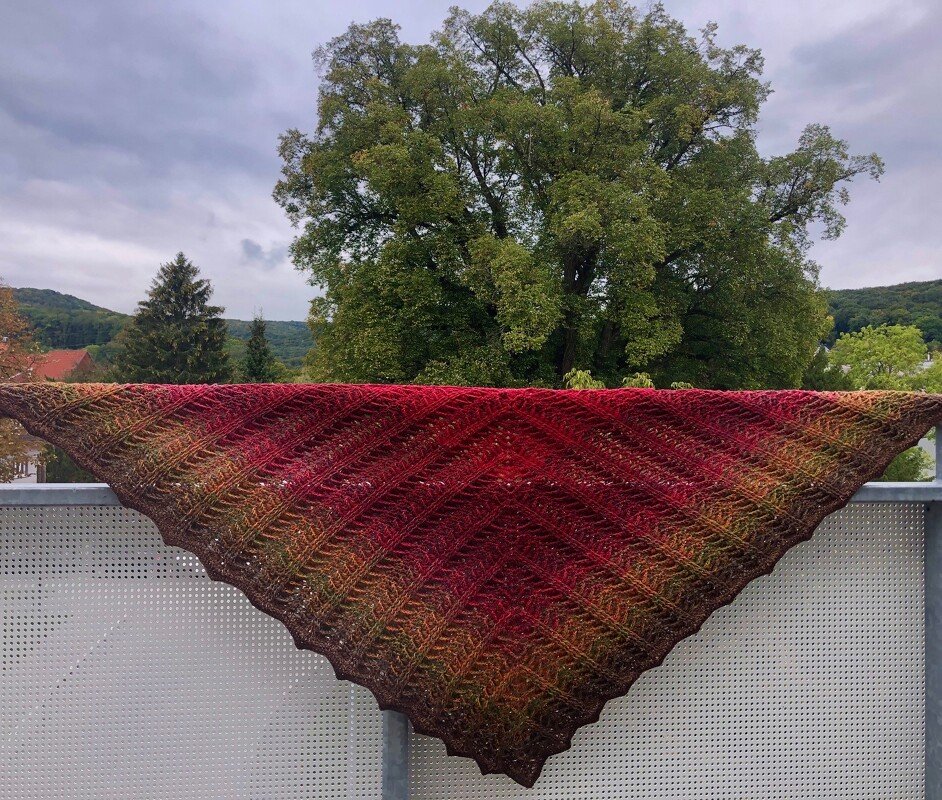 Gehäkeltes Dreieckstuch in Rot- und Grüntönen mit diagonalem Strukturmuster auf einem Balkongeländer vor Bäumen.