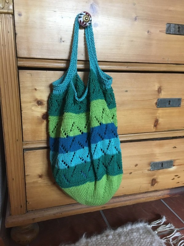 Gestrickte Einkaufstasche mit grünen und blauen Streifen und Ajourmuster, an einer Holzkommode hängend
