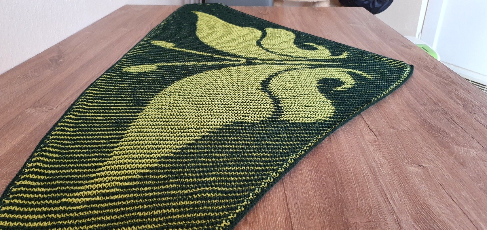 Two-tone knitted shawl with large leaf and scroll motif in dark green and lime, laid flat on a wooden table