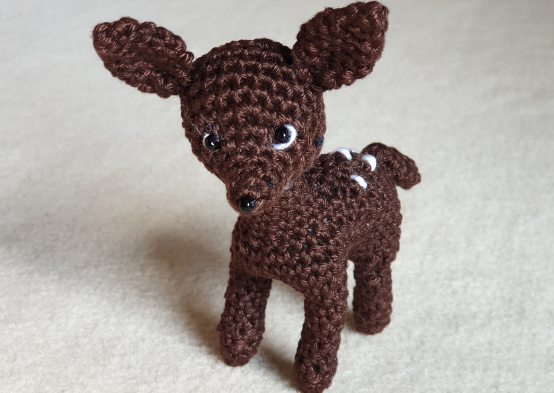 Gehäkeltes braunes Rehkitz-Amigurumi mit weißen Punkten auf dem Rücken, steht auf beigem Stoff.