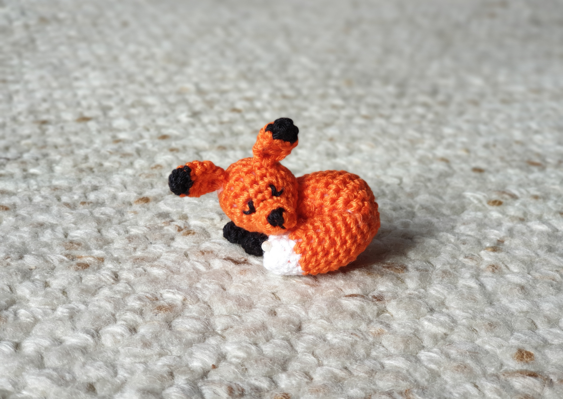 Kleiner zusammengerollter Amigurumi-Fuchs in Orange auf hellem, texturierten Untergrund