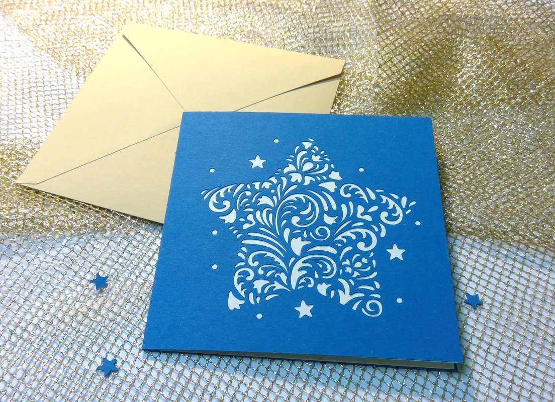 Blaue Karte mit großem, verziertem Sternausschnitt und beigefarbenem Umschlag auf goldenem Netzstoff.