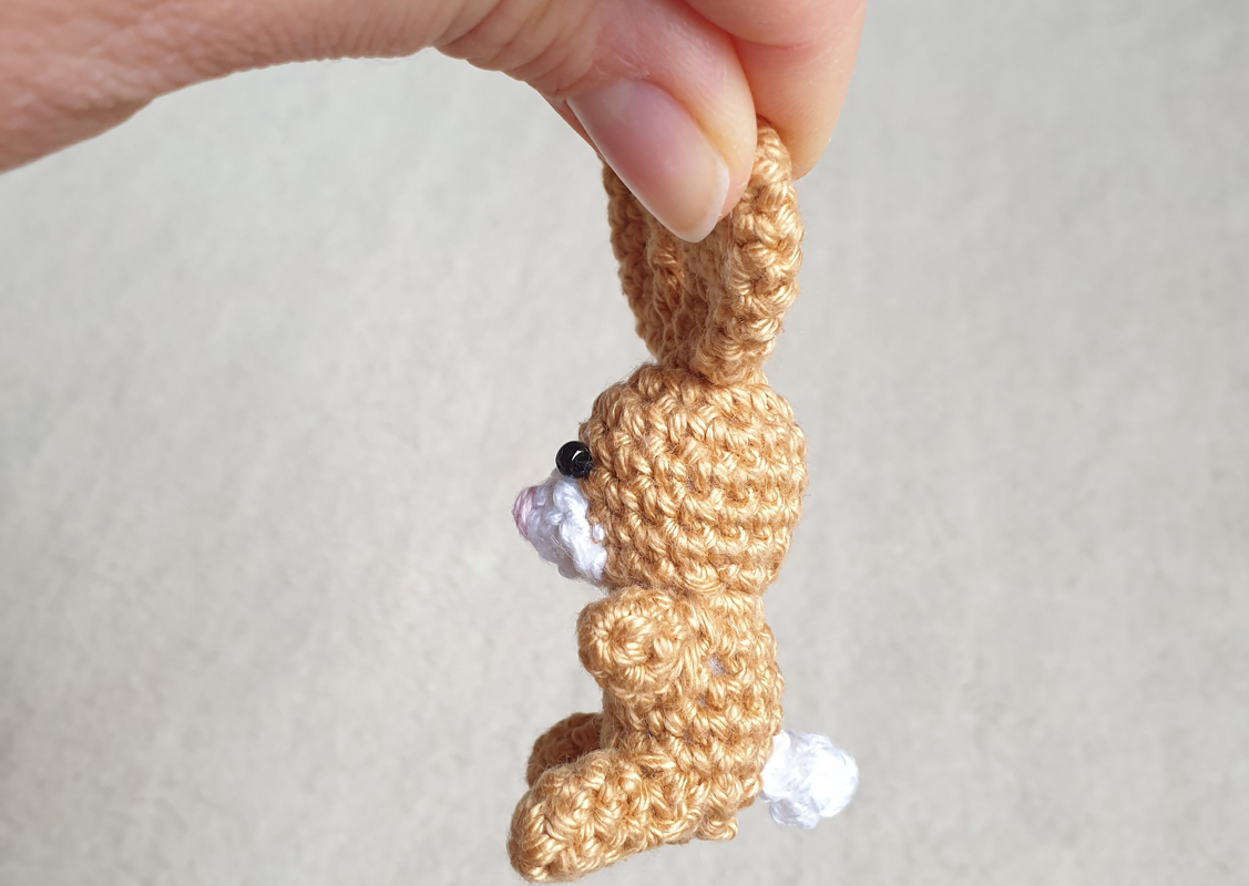 Beiger gehäkelter Mini-Hase mit weißem Schwänzchen, Seitenansicht, an Ohrspitze gehalten