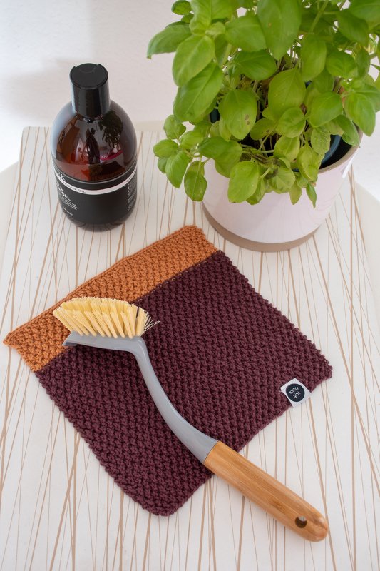 Zweifarbiges gestricktes Spültuch in Dunkelrot und Ocker mit Holzgriff-Bürste, brauner Seifenflasche und Topfpflanze auf hellem Untergrund
