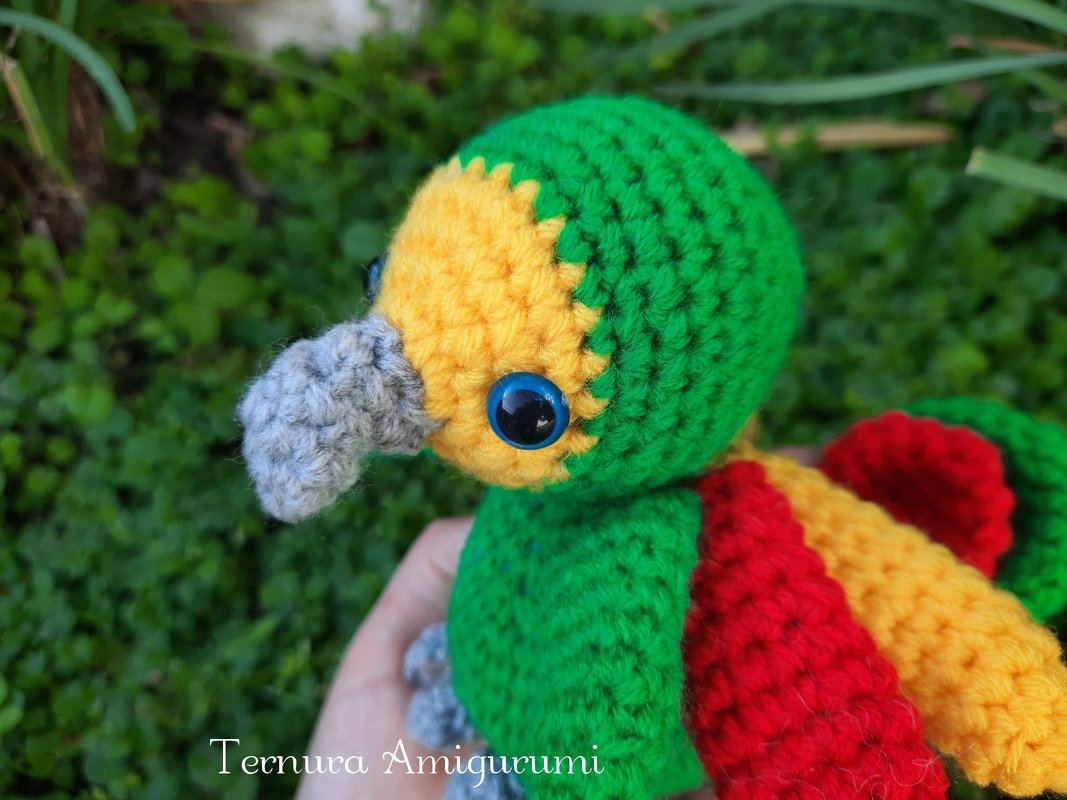 Close-up of a green, yellow and red crocheted amigurumi parrot with a grey beak and blue eyes held in a hand.