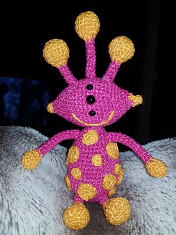 gehäkeltes Amigurumi‑Alien in Pink und Gelb mit drei Antennen und schwarzen Perlenaugen