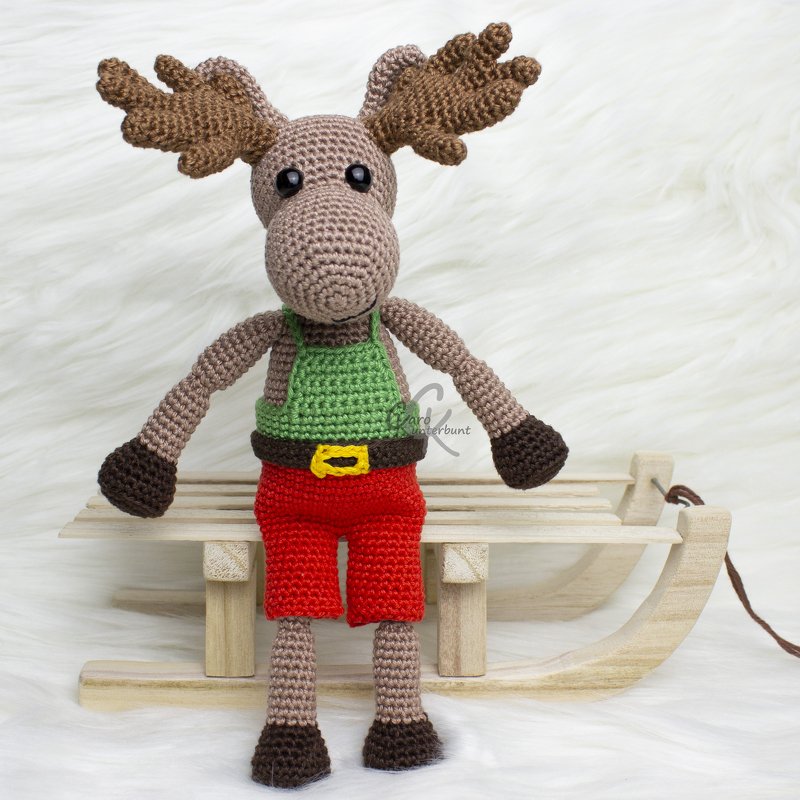 Handgearbeitetes, gehäkeltes Elch-Amigurumi mit braunem Geweih, grünem Oberteil, roter Hose und dunklen Füßen auf hölzernem Dekoschlitten